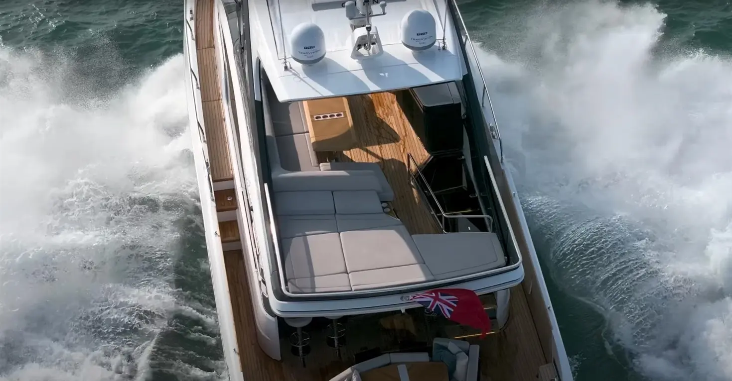 Aerial view of a luxury motorboat with seating area and Union Jack flag, creating waves in blue water.